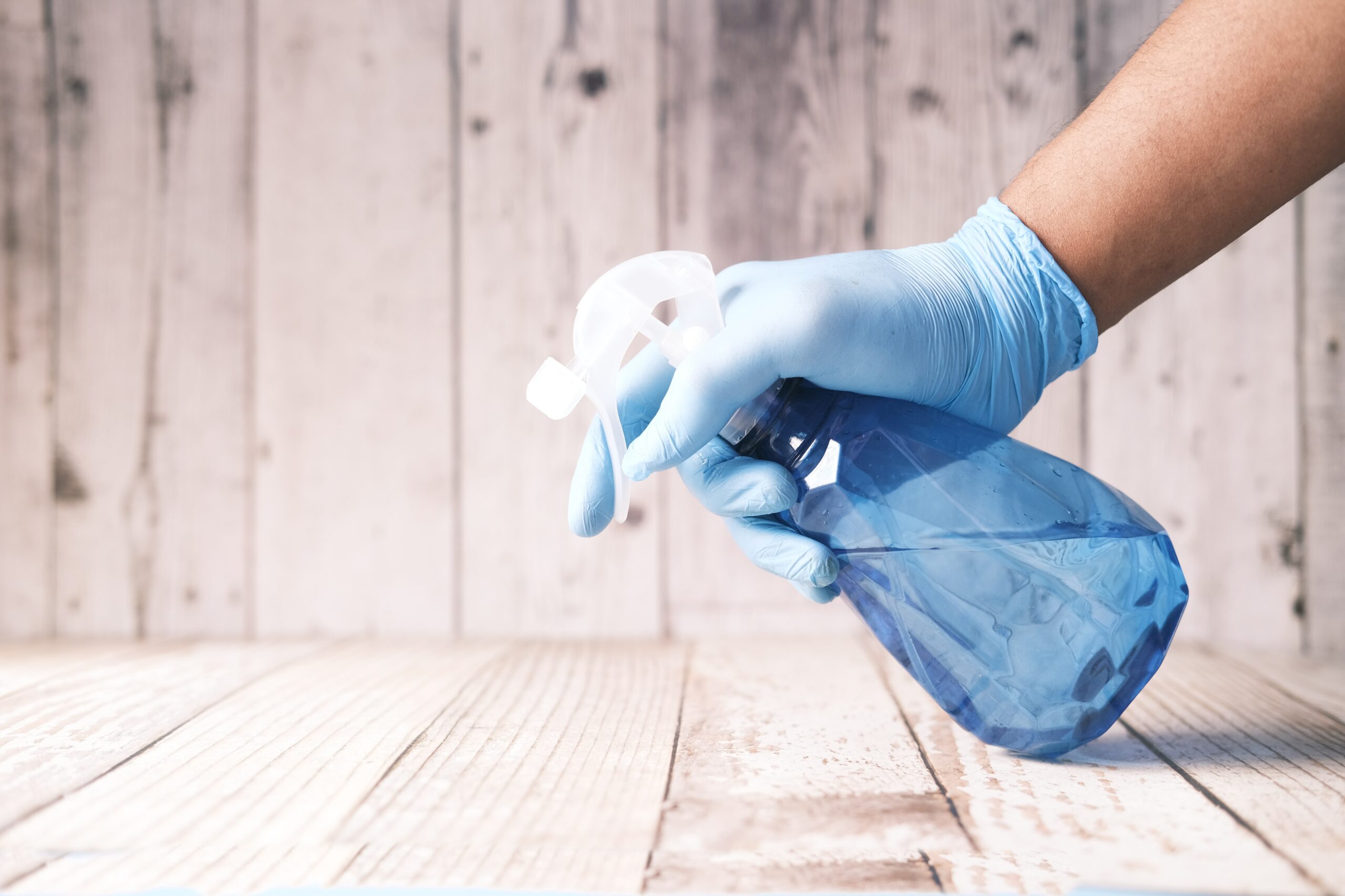 A hand wearing a rubber glove and holding a bottle of cleaning fluid over a surface as part of a side job for real estate agents